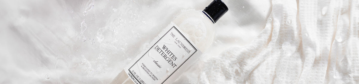 The Laundress Whites Detergent formulated to whiten & brighten laundry laying in water with white fabric.